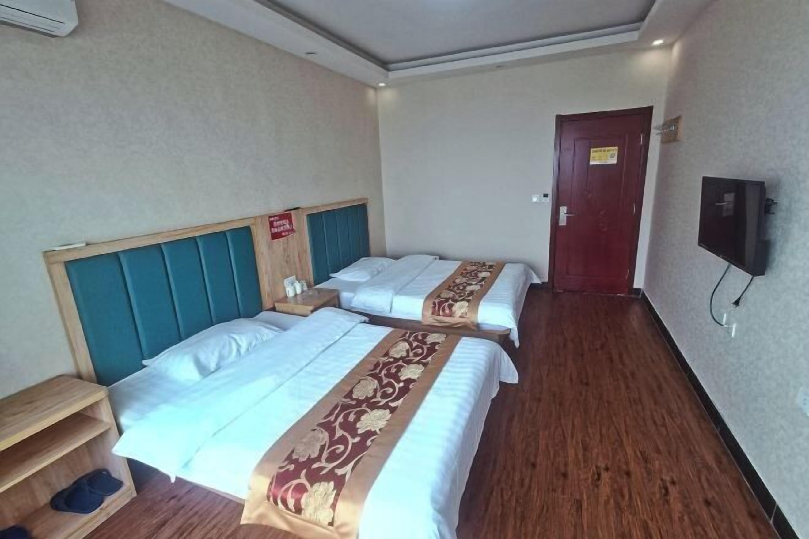 Une chambre d’hôtel avec deux lits, une table de chevet en bois, une étagère avec des chaussons, une télévision et une porte avec une pancarte.
