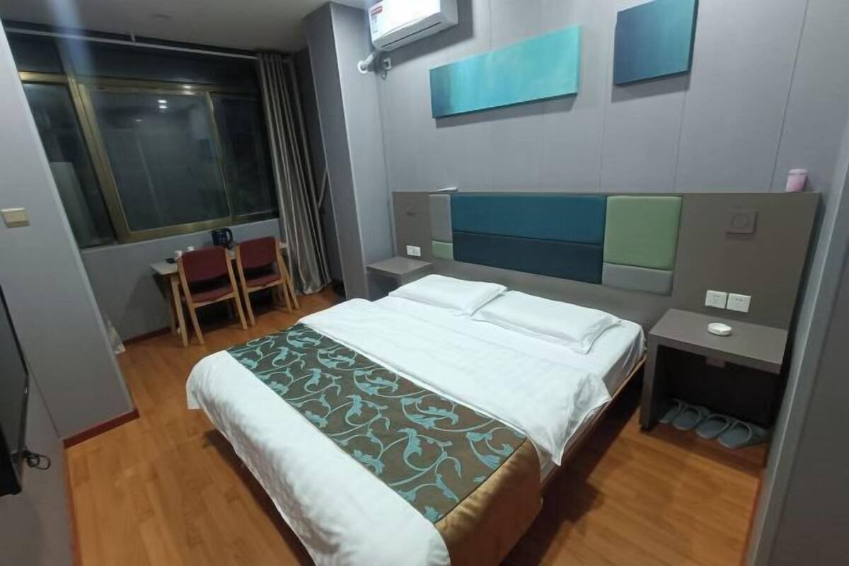 A hotel room with a bed, a desk, a chair, a window, and a shoe rack.