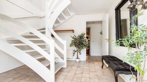 Una escalera con barandillas blancas que sube a un piso superior, una planta en maceta y un banco negro en un pasillo bien iluminado con suelo de baldosas.
