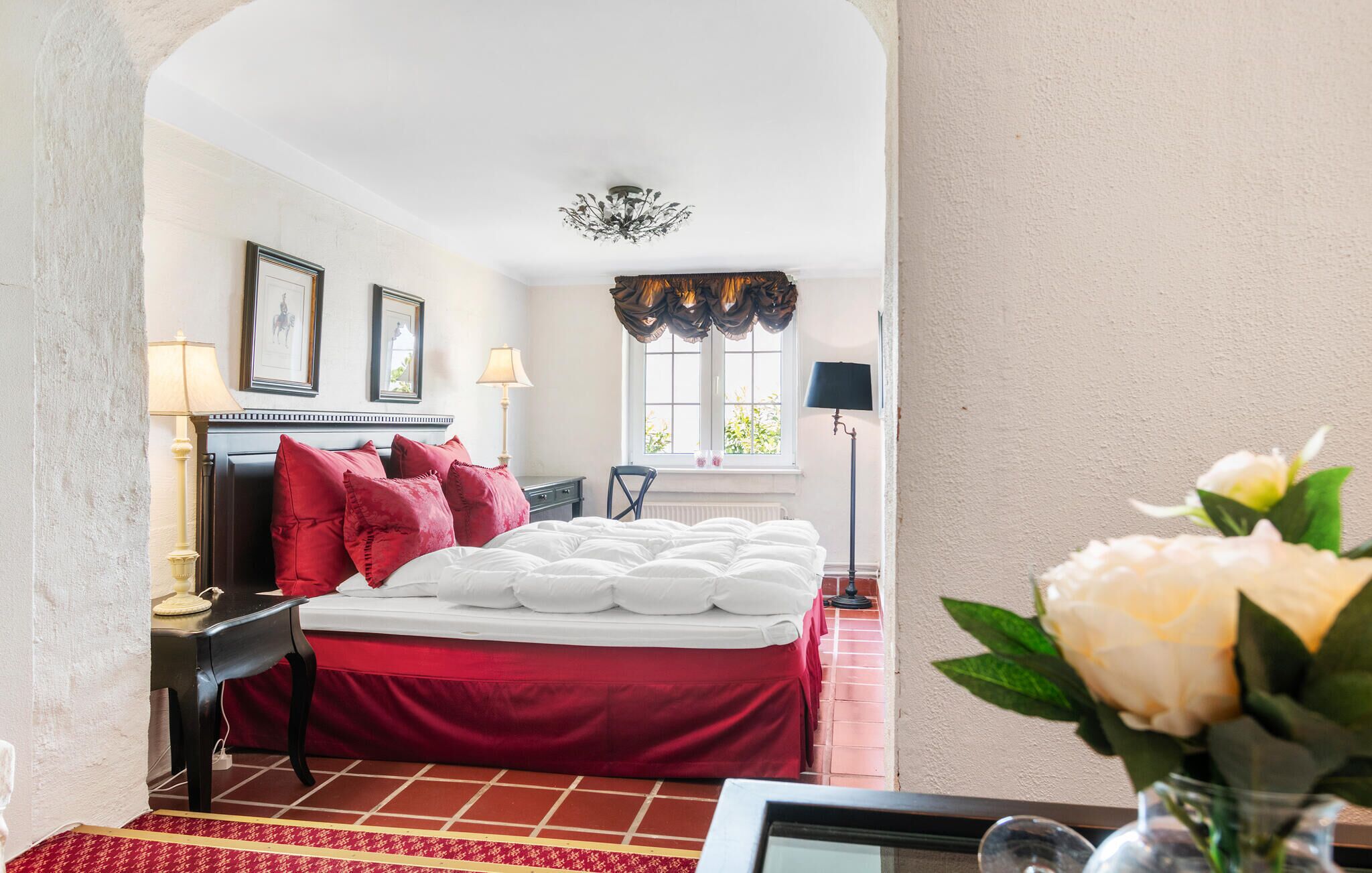 Une chambre d’hôtel comprenant un lit, deux tables de chevet, une chaise, une fenêtre avec des rideaux, un lustre et un vase avec des fleurs.