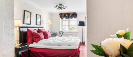 Une chambre d’hôtel comprenant un lit, deux tables de chevet, une chaise, une fenêtre avec des rideaux, un lustre et un vase avec des fleurs.