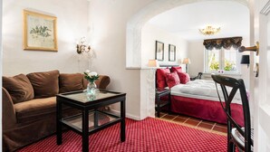 Una habitación con un sofá marrón, una mesa de vidrio con un florero, alfombra roja estampada, cama con ropa de cama roja y un candelabro.