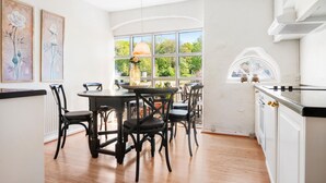 Cocina moderna con armarios blancos, zona de comedor con sillas negras y grandes ventanales con vistas a zonas verdes.