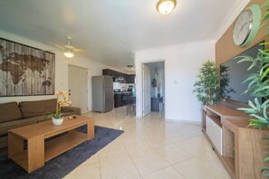 A living room with a sofa, coffee table, and a TV stand with plants. There is a kitchen in the background. - Garden Condos Apt 35 - Affordable 1br Apt In Sosua (Sosúa)