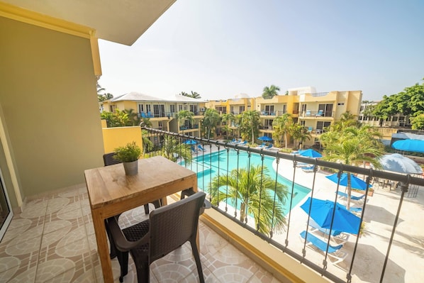 A balcony with a wooden table and chair, overlooking a swimming pool with lounge chairs and umbrellas.