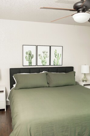 Une chambre à coucher avec un lit, une table de chevet, une lampe et trois tableaux représentant des cactus, accrochés au mur.