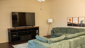 Een woonkamer met een grote flatscreen-tv, een groene bank, een vloerlamp en een muur met drie schilderijen.