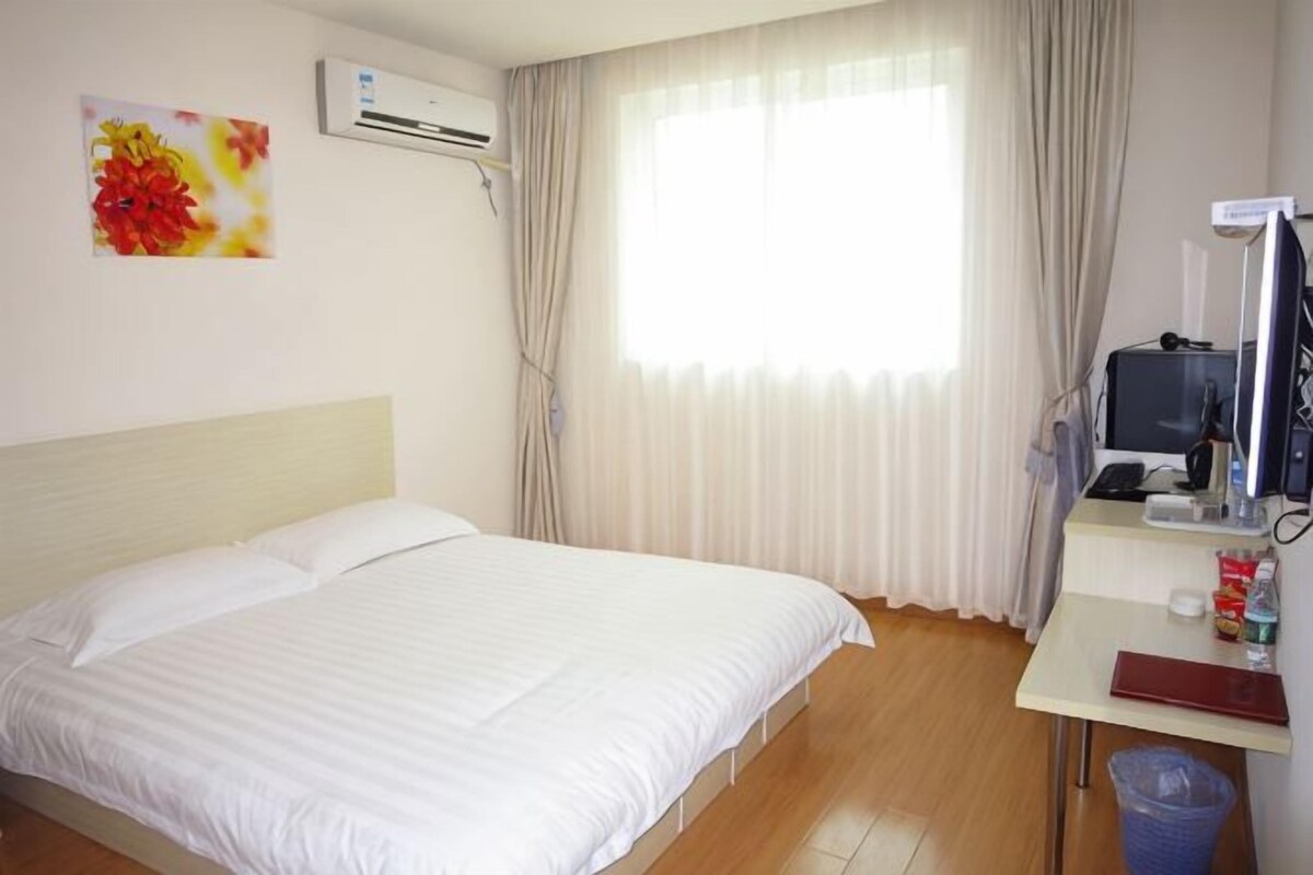 A hotel room with a bed, a desk, an air conditioning unit, a window with curtains, and a trash bin.
