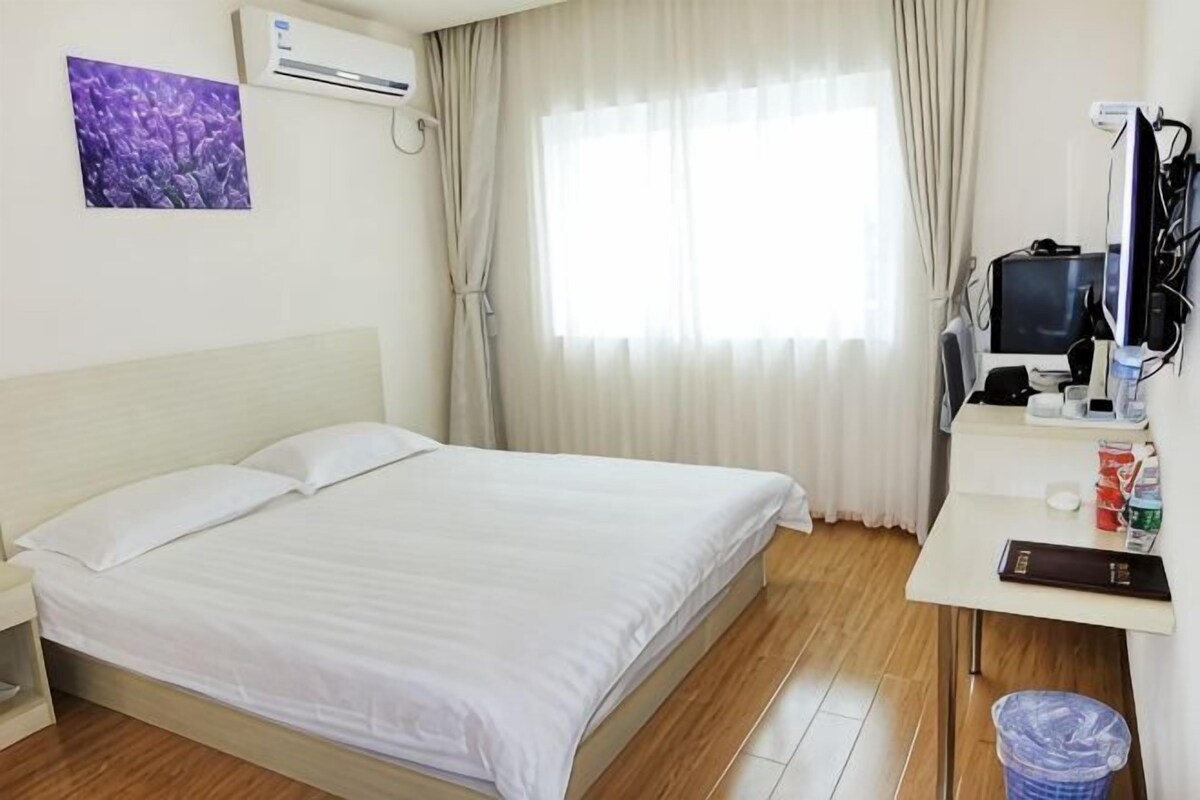 A hotel room with a bed, a desk with a computer, a trash bin, and a window with curtains.