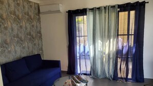 Un saló amb un sofà blau, una taula de centre de vidre amb aperitius, i una vista a un espai exterior a través de cortines translúcides.