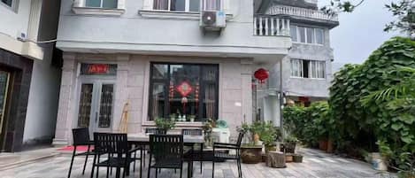 A courtyard with a dining table and chairs, surrounded by potted plants and a building with a traditional design.