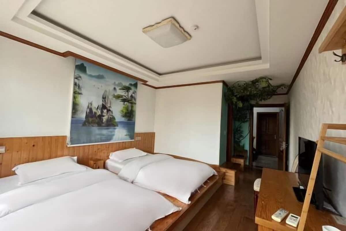 A hotel room with two beds, a ceiling light, a painting, a TV, and a wooden desk.