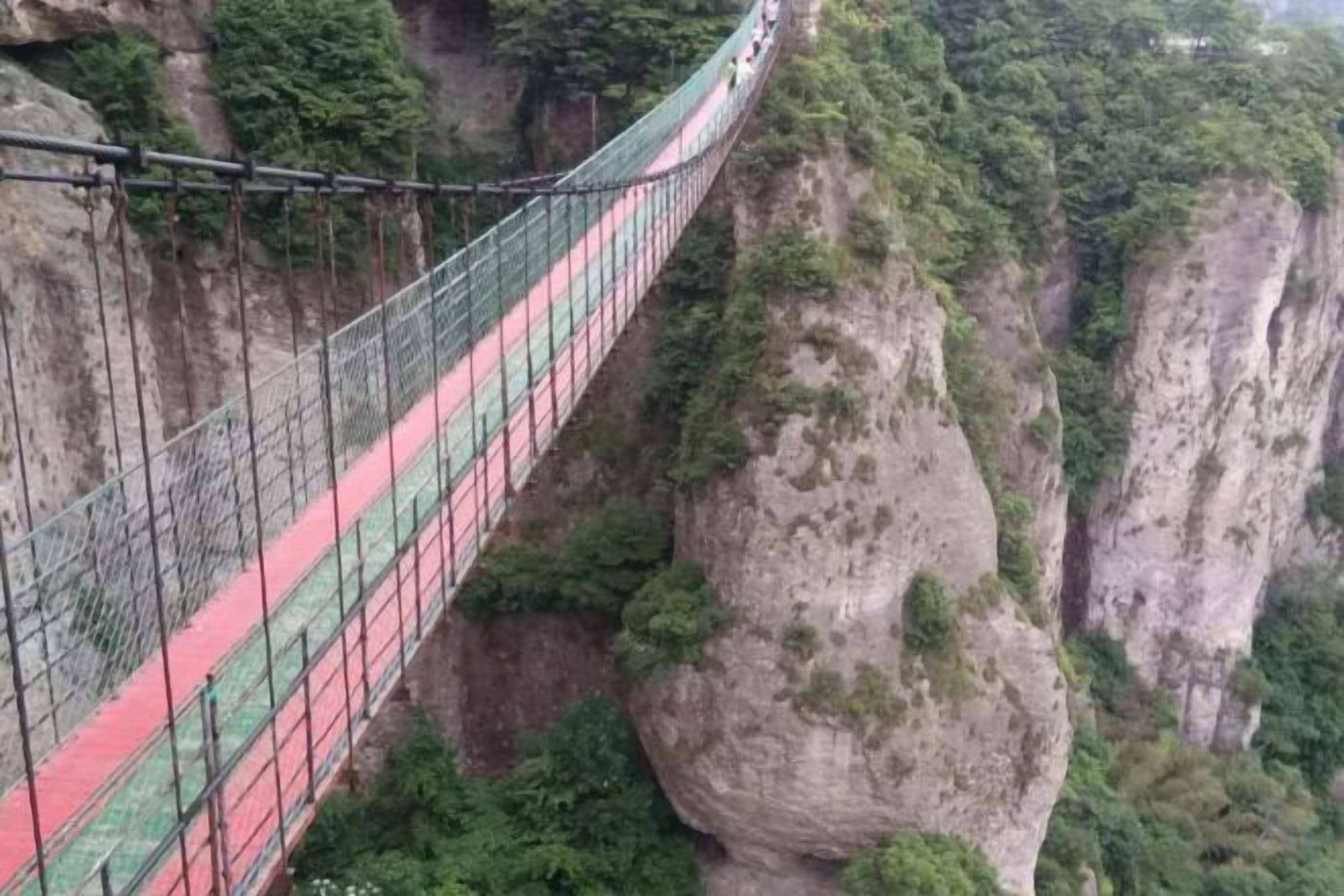Eine Hängebrücke mit rotem und grünem Gehweg, umgeben von Felsen und Grünflächen.