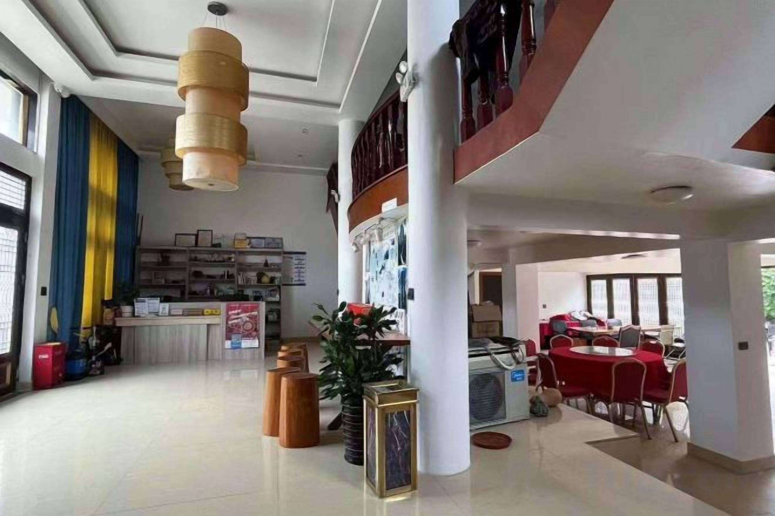 Un vestíbulo espacioso con mostrador de recepción, zona de comedor con mesas rojas y una escalera que sube a un nivel superior.