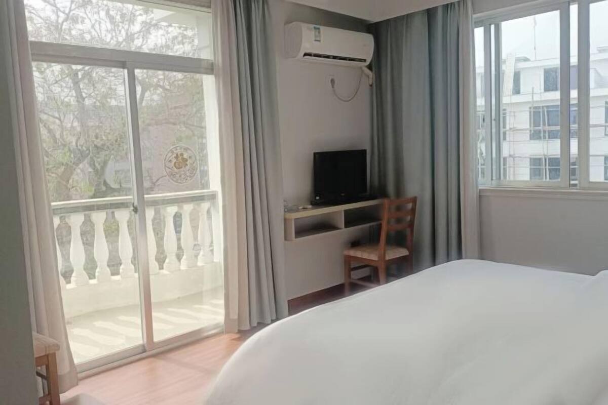 A hotel room with a bed, a desk with a television, and a balcony with a view of trees and buildings.