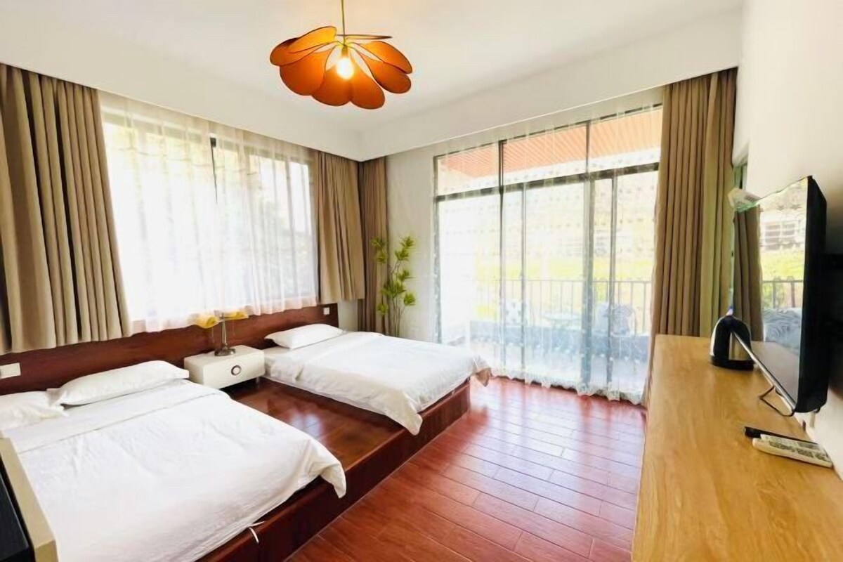 A hotel room with two beds, a wooden desk, a television, and large windows with curtains.
