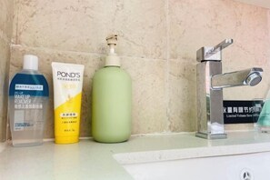 A bathroom with a sink, a mirror, and toiletries including a Maybelline makeup remover, a Ponds lotion, and a green bottle.