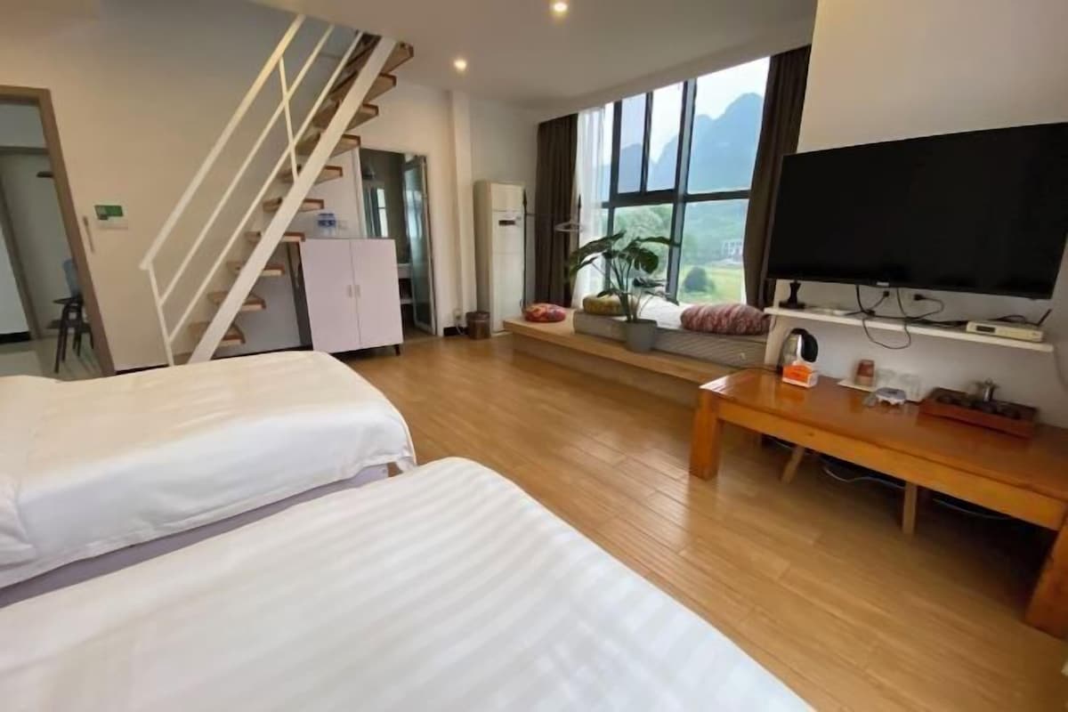 A hotel room with two beds, a TV, a desk, a staircase, and a view of the outdoors.
