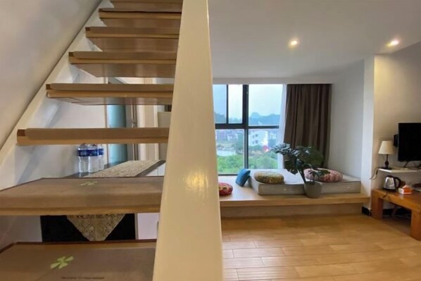 A modern living room with a wooden staircase, a large window with a view, and a cozy seating area with cushions and a plant.