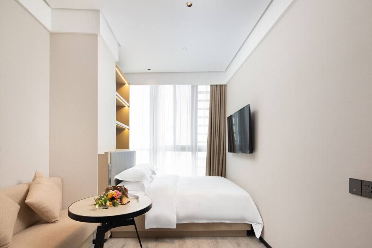 Une chambre d’hôtel avec un lit, un canapé, une petite table ronde agrémentée d’un bouquet de fleurs, un téléviseur fixé au mur et une fenêtre avec des rideaux.