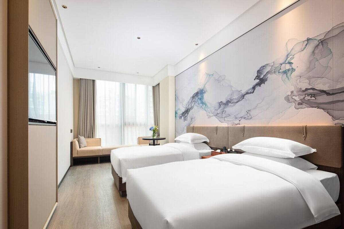 Une chambre d’hôtel avec deux lits, une grande peinture abstraite au mur, un canapé et une vue sur la ville grâce à de grandes fenêtres.