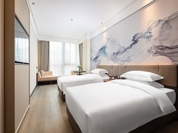 Habitación de hotel con dos camas, un cuadro abstracto grande en la pared, un sofá y vistas de la ciudad a través de ventanas amplias.