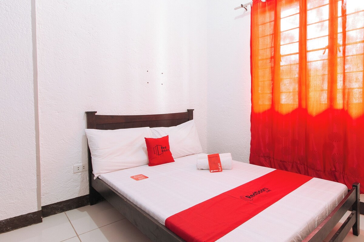 A neatly made bed with a red bedspread and pillows, a wooden headboard, and a window with orange curtains.