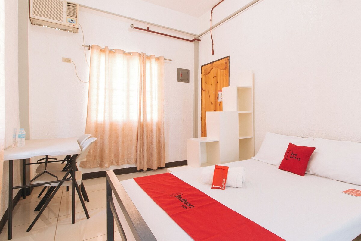 A hotel room with a bed, a desk, and a chair. There is a window with curtains, a wall-mounted shelf, and a door.