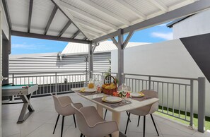 Un espace extérieur abrité, aménagé pour les repas, avec une table de billard, des chaises et une table dressée pour deux personnes, avec des assiettes, des verres et un panier de fruits.