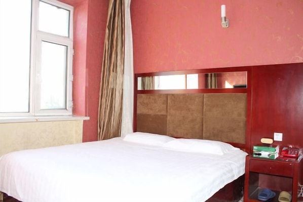 Una habitación con cama, ventana, espejo y mesita de noche con teléfono y una caja.