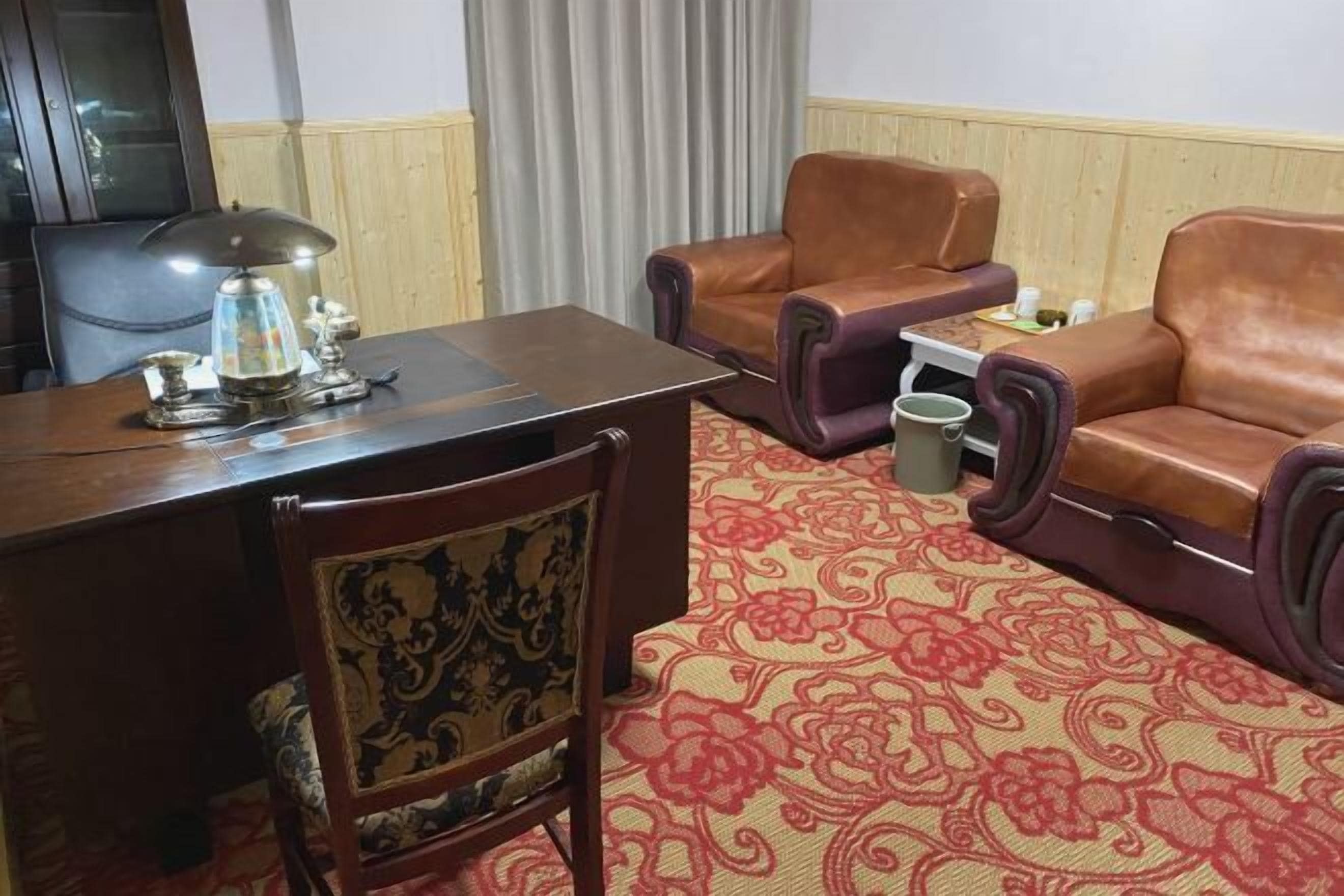 A room with a wooden desk, a lamp, a patterned carpet, leather armchairs, and a small table with a trash bin.