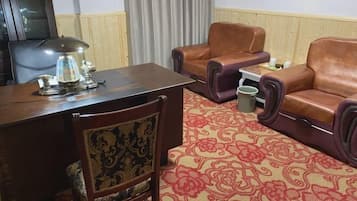 A room with a wooden desk, a lamp, a patterned carpet, leather armchairs, and a small table with a trash bin.