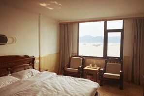 Habitación de hotel con una cama grande, dos sillones, una mesita y una ventana con vistas al agua y montañas.