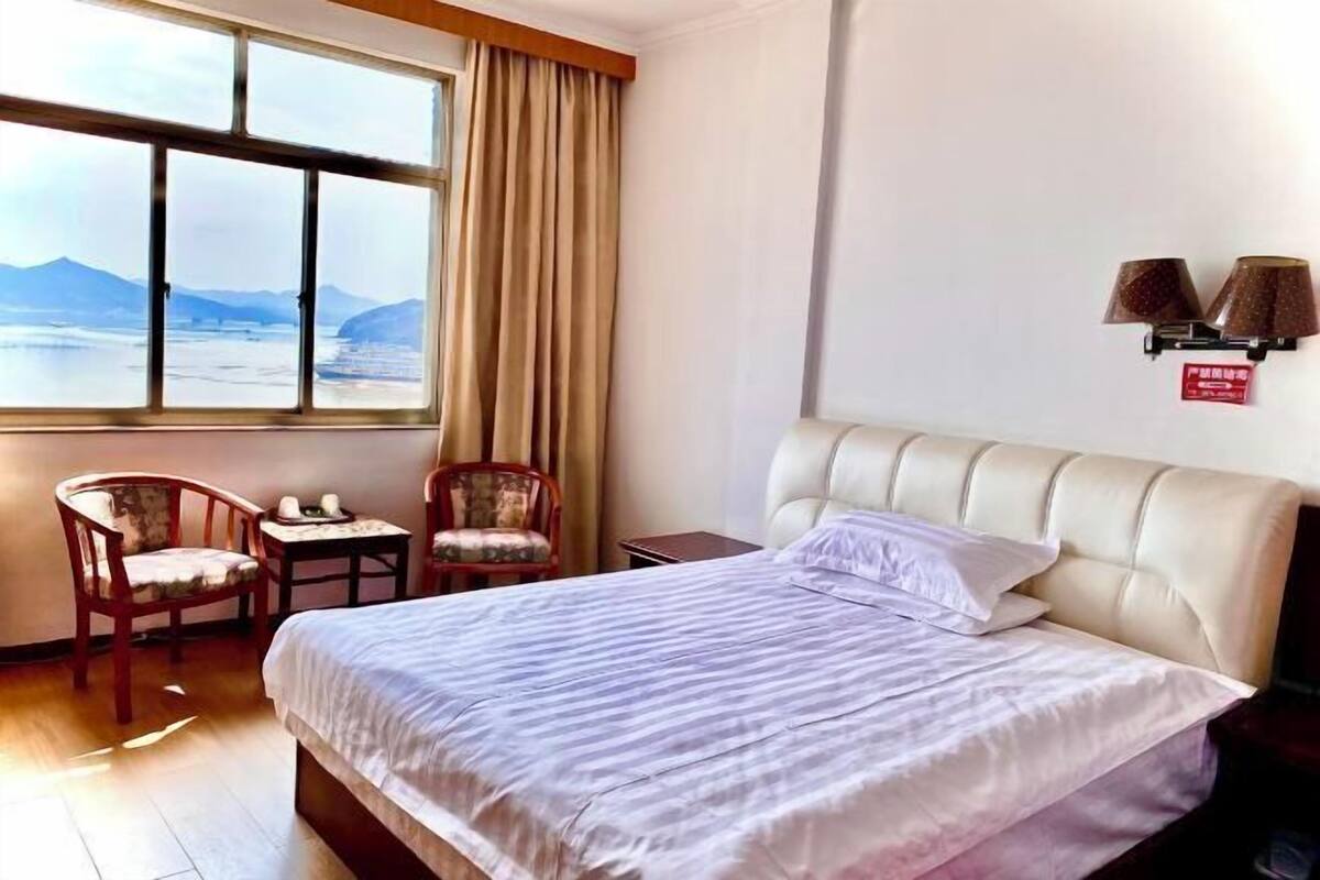 A hotel room with a large bed, two chairs, a small table, a window with a view of mountains, and a wall-mounted lamp.