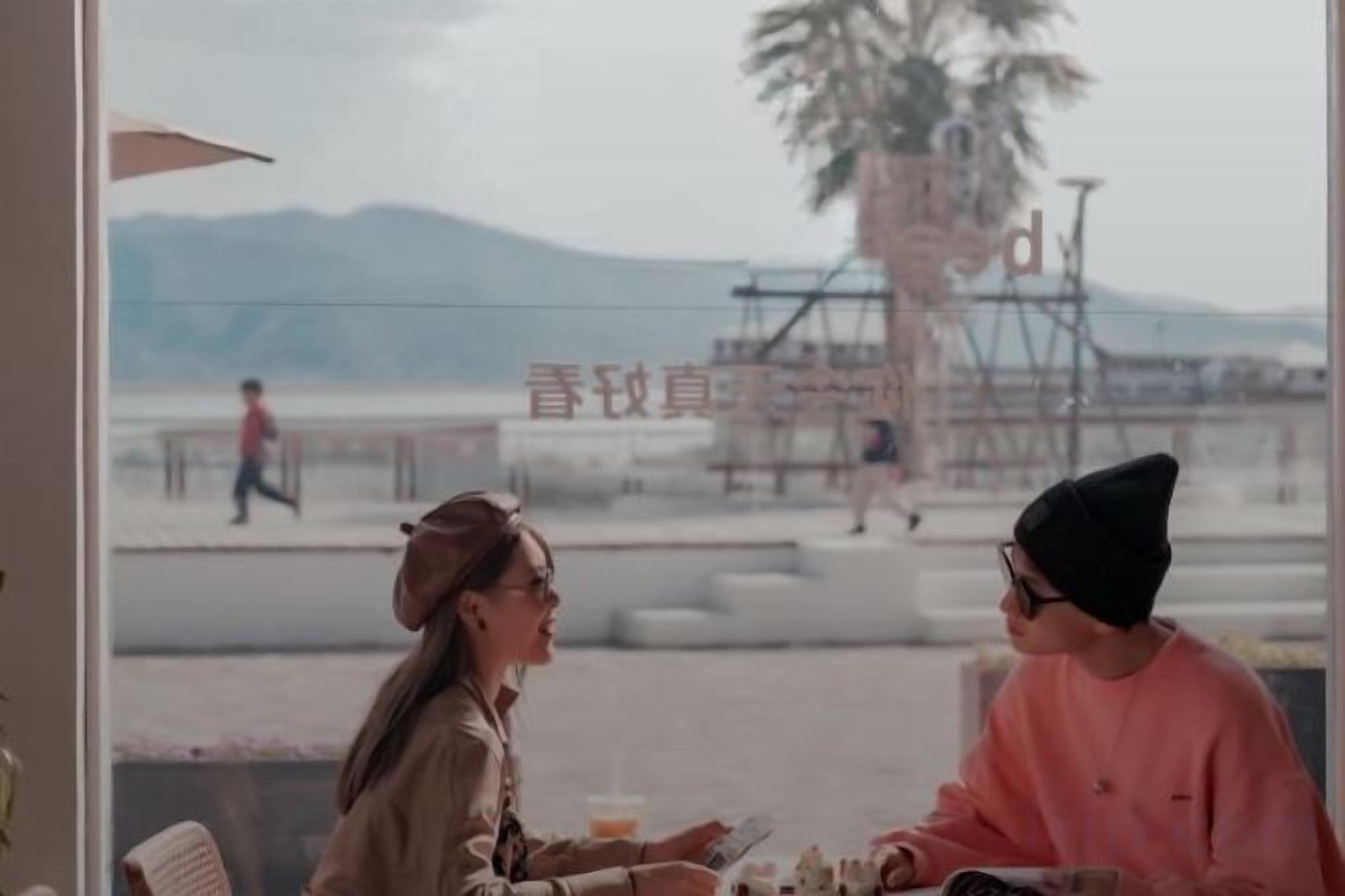 Una persona sentada a una mesa con una bebida, otra persona de pie al fondo, y una vista a una playa y montañas a través de una ventana.