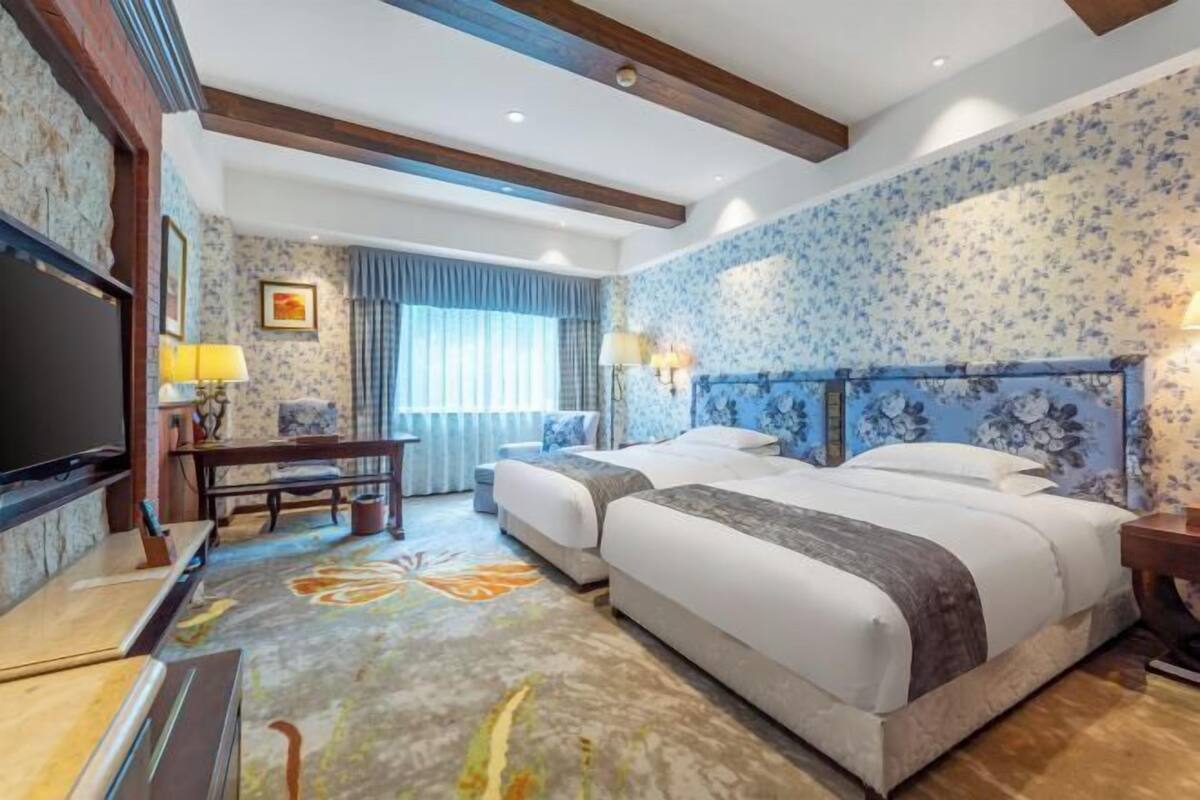 A hotel room with two beds, a flat-screen TV, a desk, and a large window with curtains.
