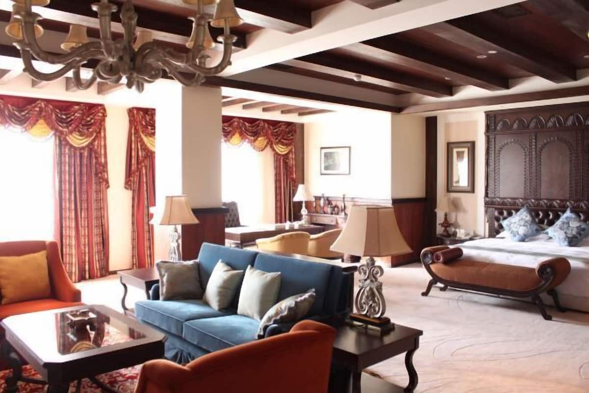 A hotel room with a bed, a blue sofa, an orange armchair, a wooden bench, and a chandelier.