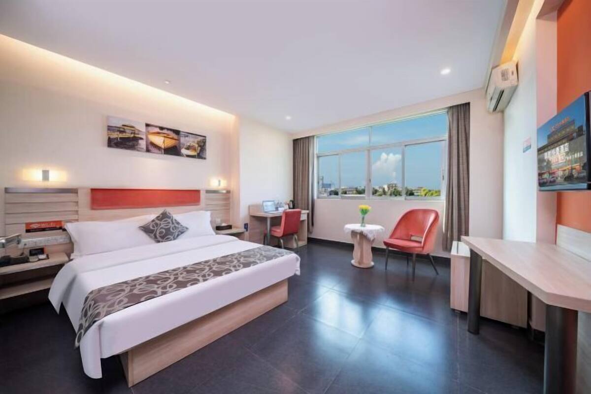 A modern hotel room with a large bed, a desk, a chair, and a small table. There is a window with a view of the city.