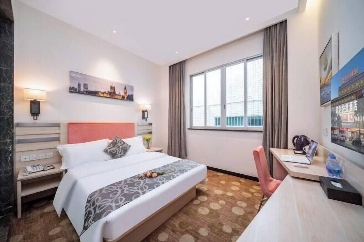 A hotel room with a bed, a desk, a chair, a window, and a cityscape painting on the wall.