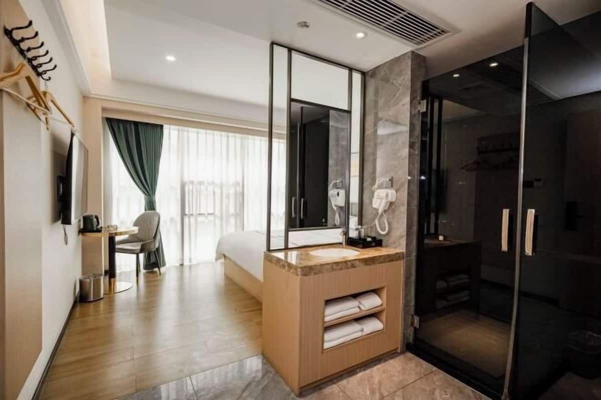 A modern hotel room with a wooden floor, a large bed, a bathroom with a glass shower enclosure, and a desk with a chair.