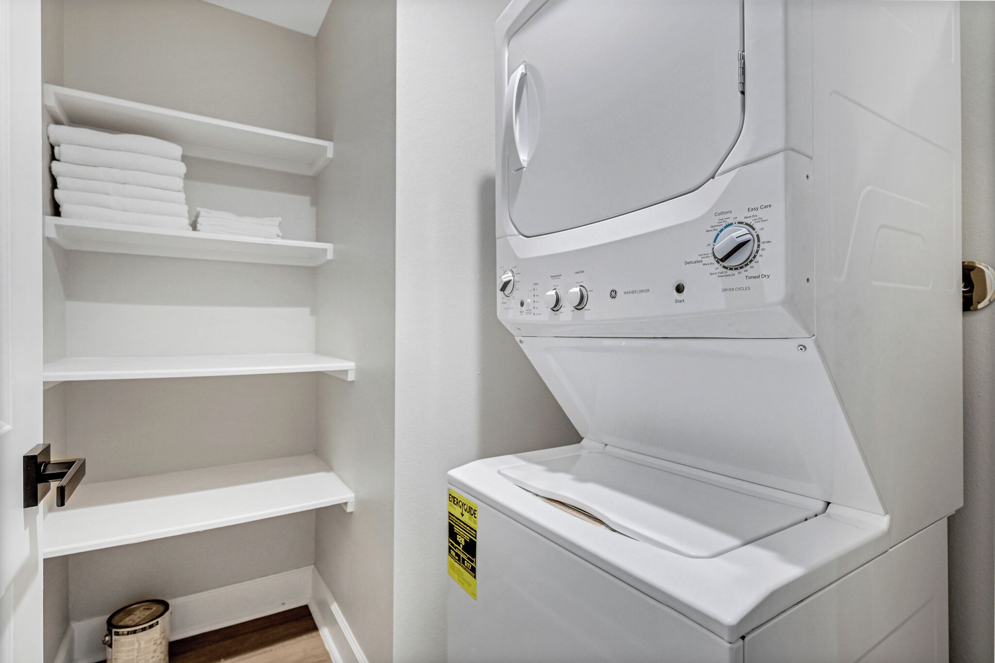 A laundry room with a front-loading washer and dryer, shelves with folded towels, and a caution sign on the dryer.