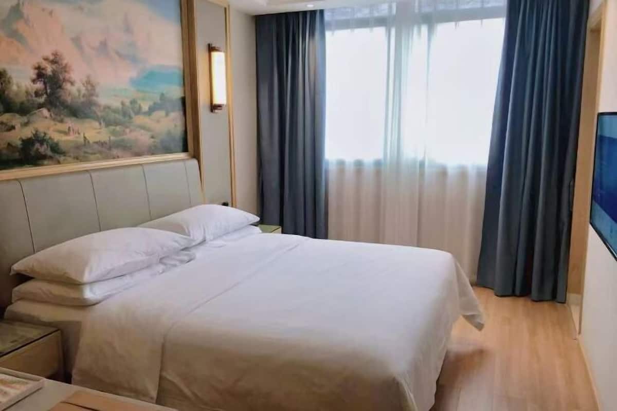 Una habitación de hotel con una cama, una mesita de noche, un cuadro en la pared y una ventana con cortinas.