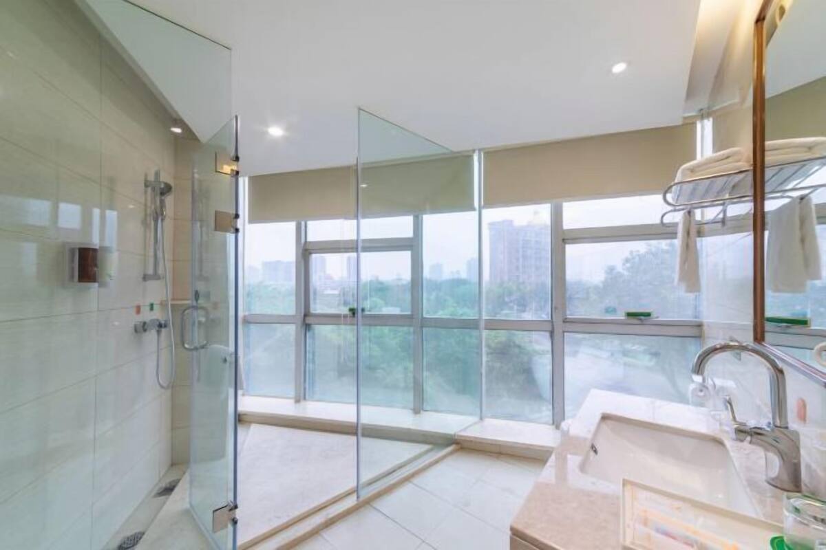 Un baño moderno con cabina de ducha de vidrio, un amplio ventanal con vistas a la ciudad y un lavamanos empotrado con toallero.