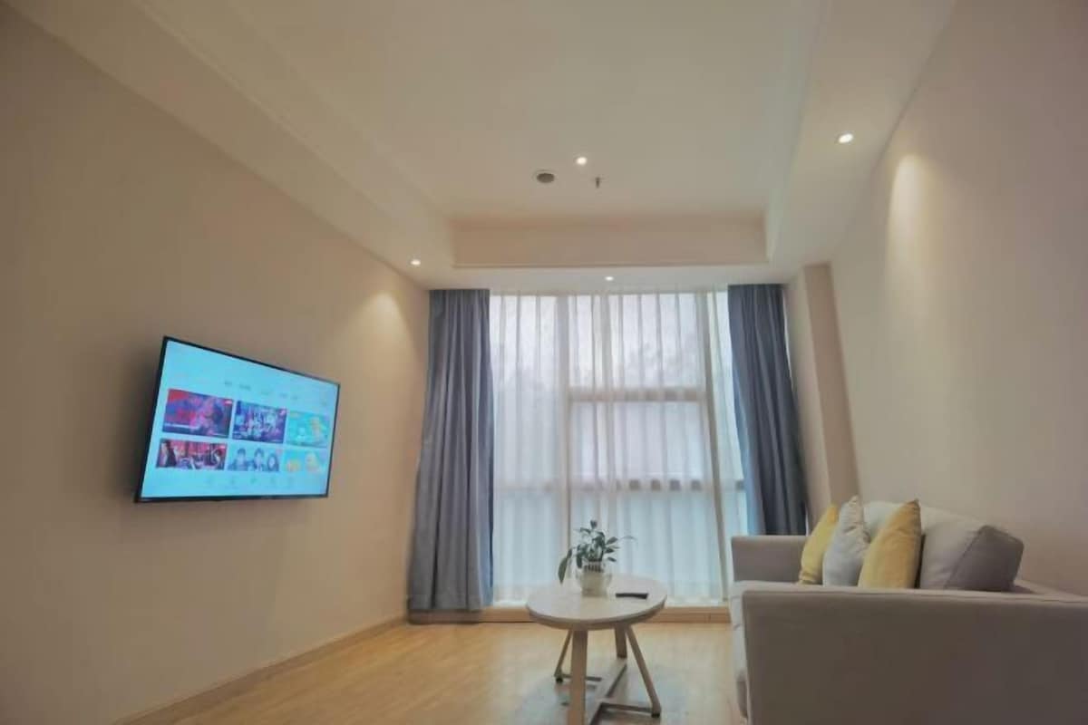 A modern living room with a flat-screen TV mounted on the wall, a round table with a plant, a sofa with cushions, and a large window with sheer curtains.