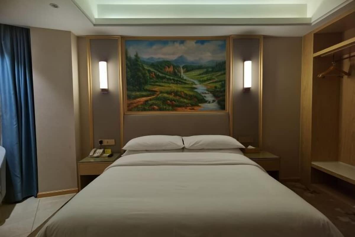 A hotel room with a large bed, bedside tables, a painting of a landscape, and a wardrobe.