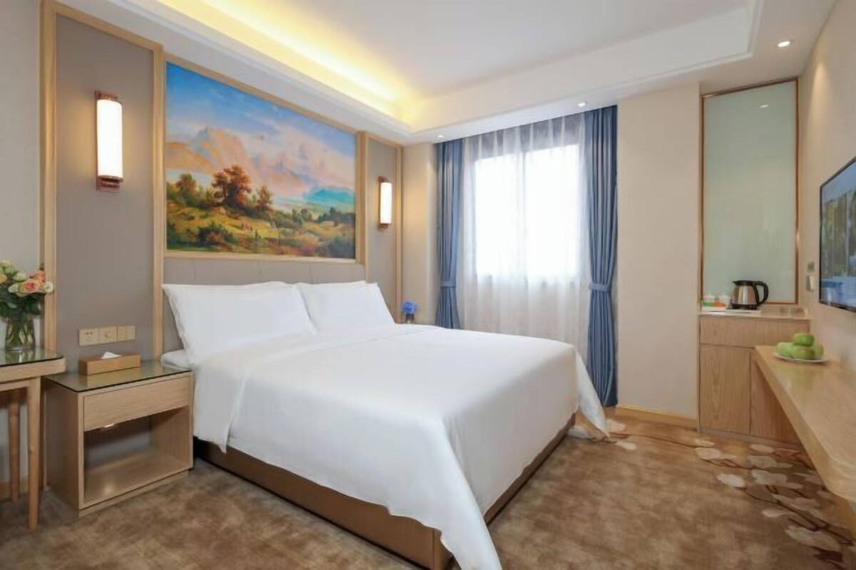 Habitación de hotel con una cama grande, un cuadro en la pared, un escritorio con una lámpara y una ventana con cortinas.