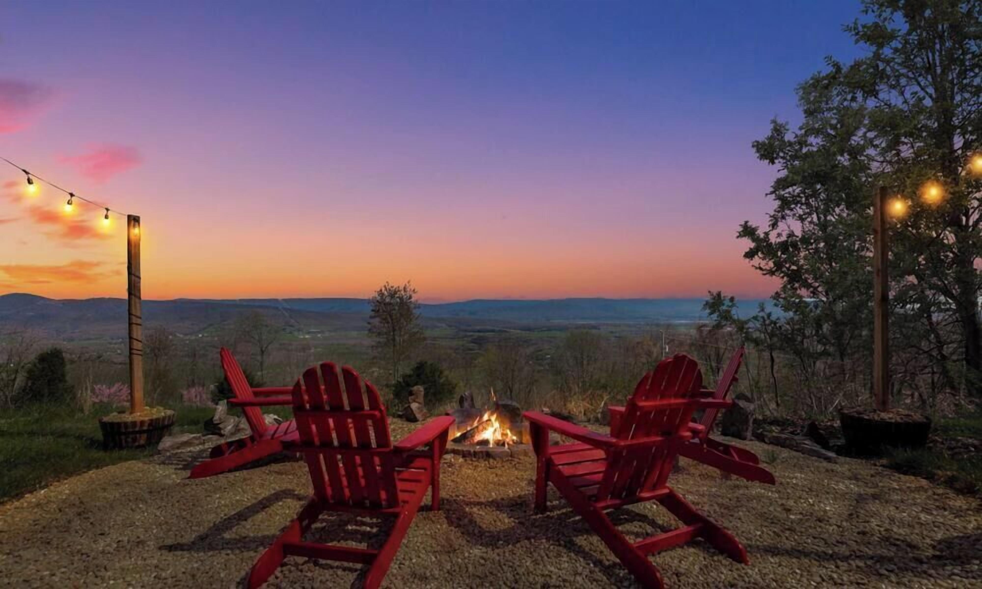 Two red Adirondack chairs facing a fire pit with a view of a sunset over a hilly landscape.
