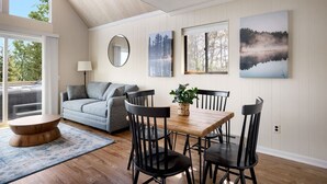Un salon chaleureux avec un canapé gris, une table basse en bois et une table à manger avec des chaises noires. Deux tableaux encadrés ornent le mur, et un miroir rond est accroché au-dessus du canapé.