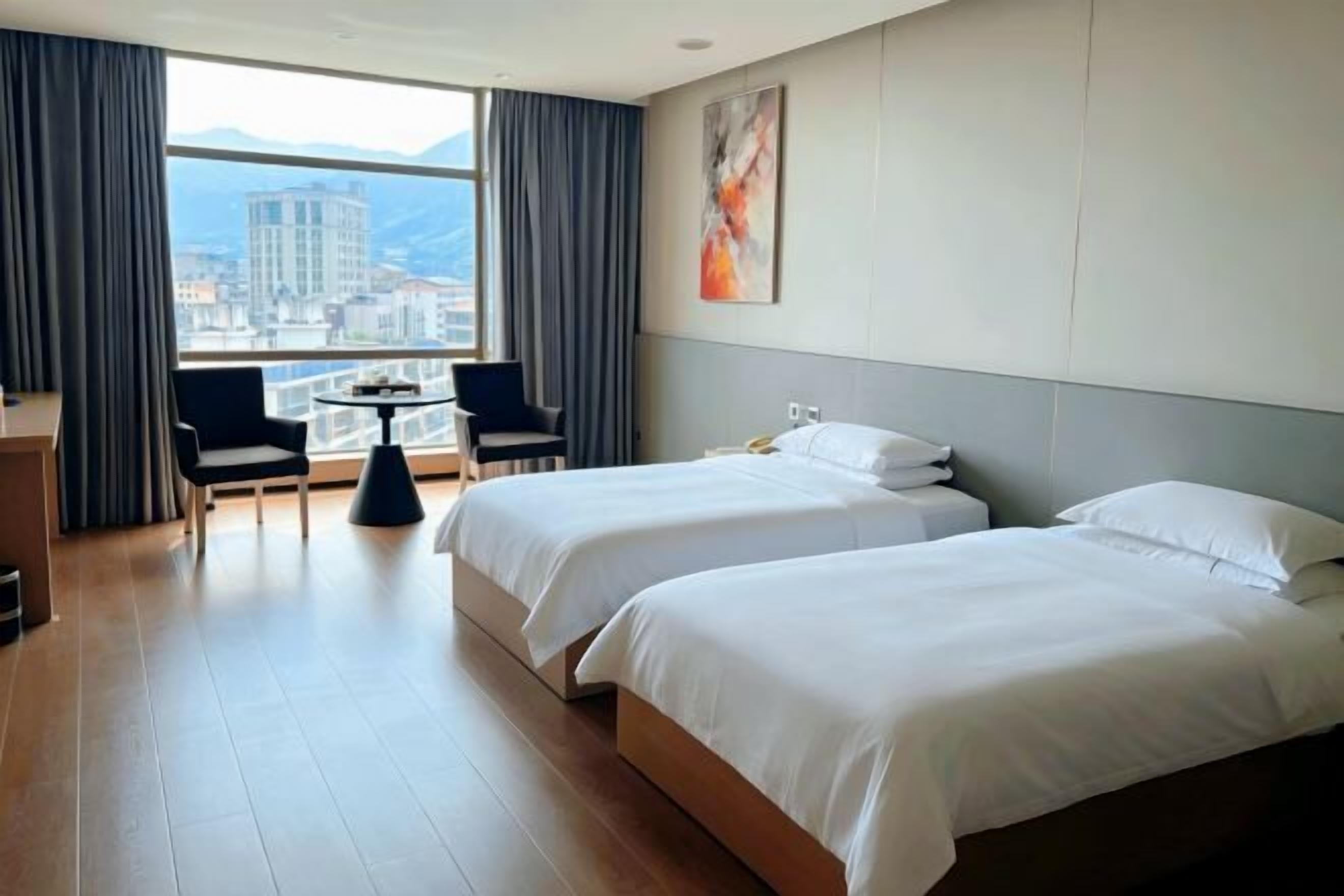 Habitación de hotel con dos camas, un escritorio, dos sillas, una mesita pequeña y un ventanal grande con vistas a la ciudad.