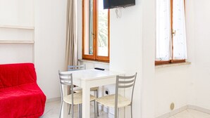 Una habitación con una mesa blanca, cuatro sillas, un sofá rojo, un televisor fijado a la pared y dos ventanas con cortinas.